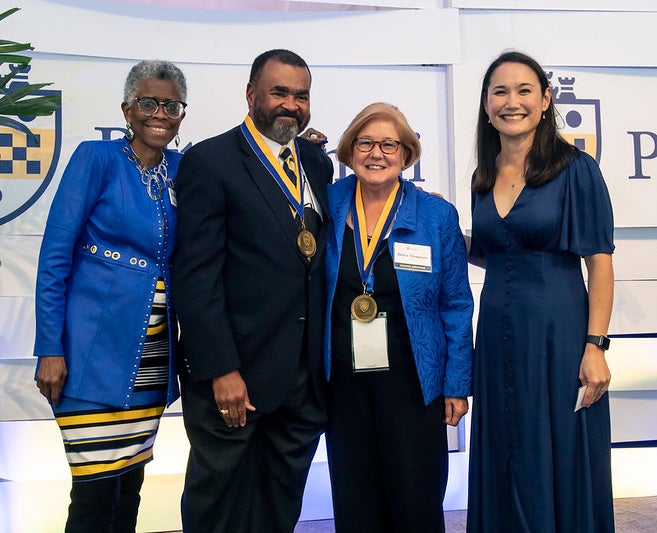 Black woman in blue, white and gold dress; Black man in suit, tie and gold medallion; white woman in blue jacket and black blouse, slacks and gold medallion; and white woman in navy blue dress pose for camera in front of Pitt-seal branded background