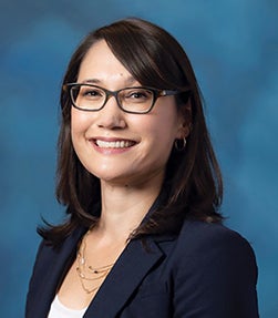 Woman with dark straight hair, dark-rimmed glasses, navy blue blazer, white shirt and layered necklaces smiles at camera in front of teal background