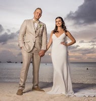 A person in a tan suit and another in a white wedding dress pose on a beach