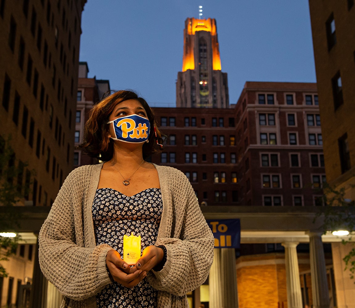 Student wearing tan cardigan and floral dress holds her LED-powered lantern and wears her Pitt script mask in the Quad, the Cathedral lights on in the background