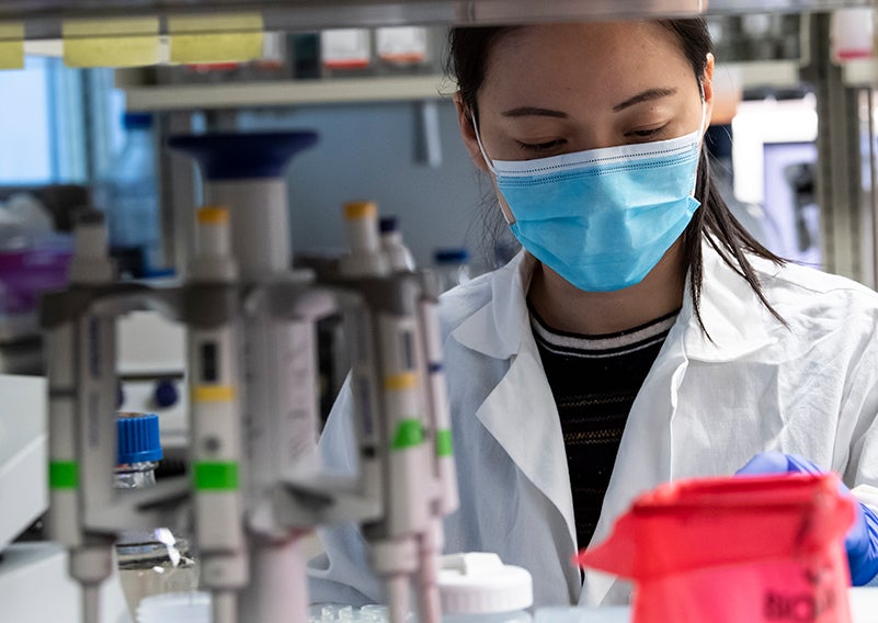 masked Asian woman wearing white lab coat seen behind open lab shelf