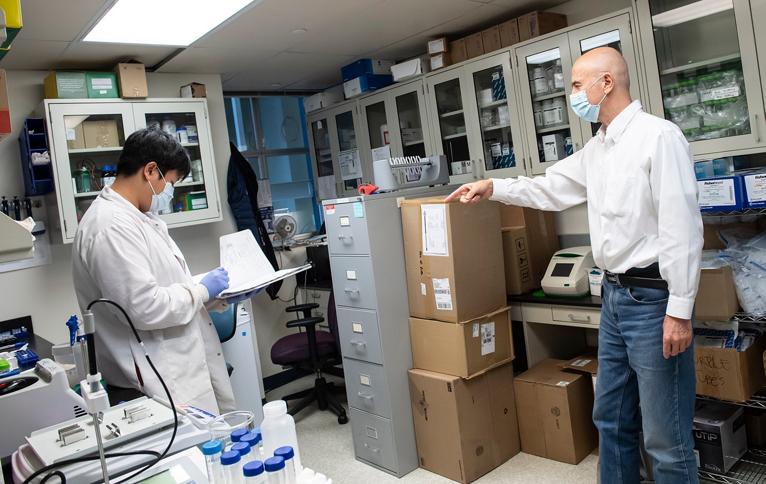 two men, one wearing blue gloves and white lab coat and holding open binder, the other in a long-sleeve white button-down tucked into jeans, stand apart and masked in lab space crowded with equipment, boxes, a desk and chair, and a filing cabinet.