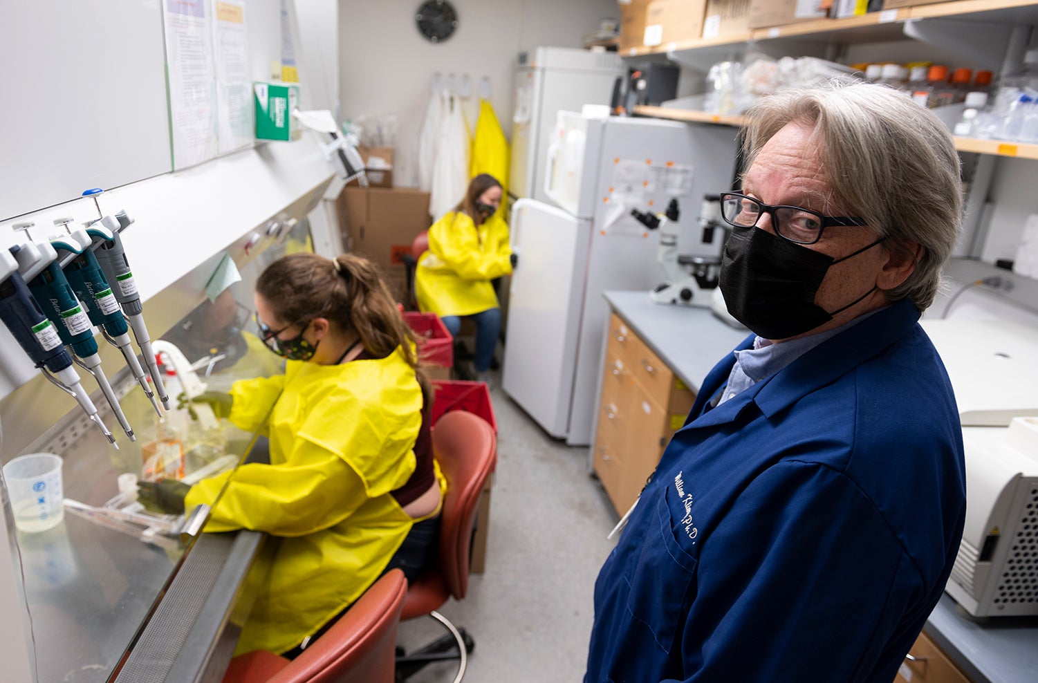 white man wearing glasses, mask, and Navy blue lab coat stands in lab and looks at camera while two women wearing masks, black gloves and yellow protective smocks work in background.