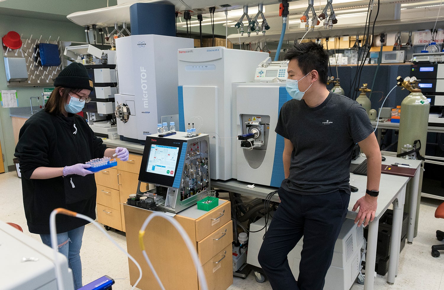 Woman wearing beanie, glasses, blue mask, dark sweater, jeans and purple gloves organizes small vials of samples while a man wearing a blue mask, dark gray short-sleeve shirt, and dark pants stands and looks on in the lab. 