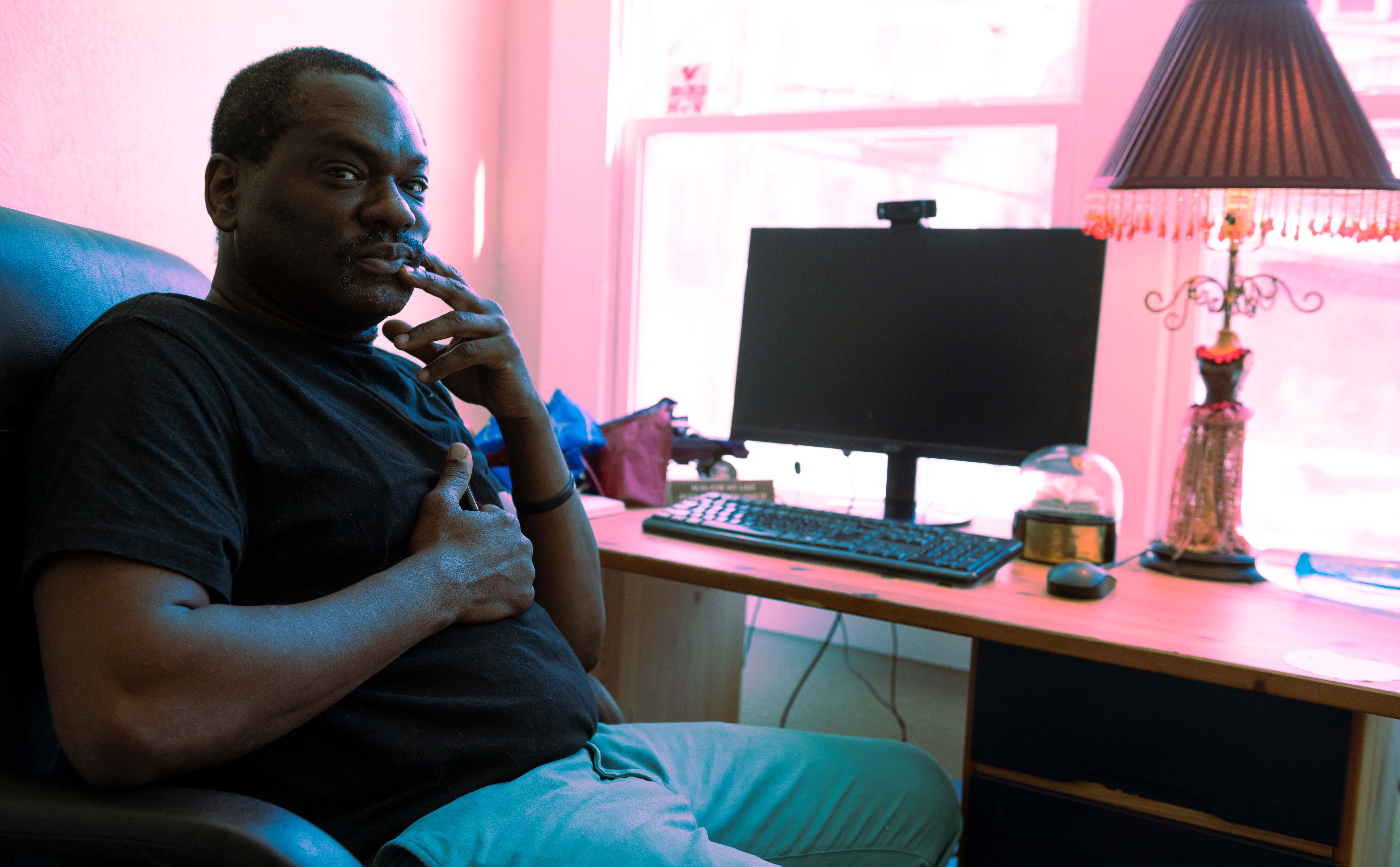 Black man in black t-shirt and turquoise slacks sits at a desk in front of a window, with a computer monitor, keyboard and table lap 