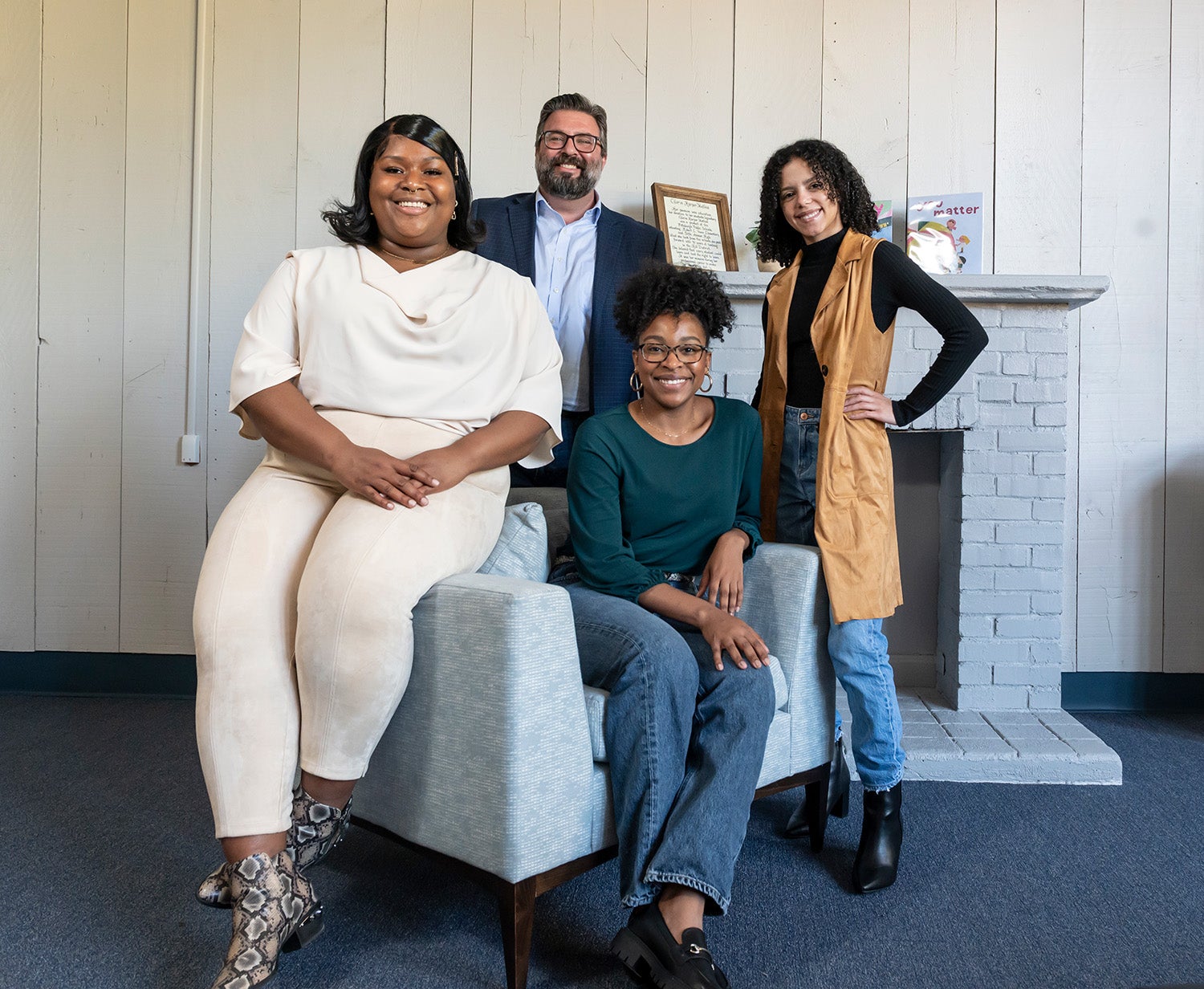 Four people sit in and stand around a chair