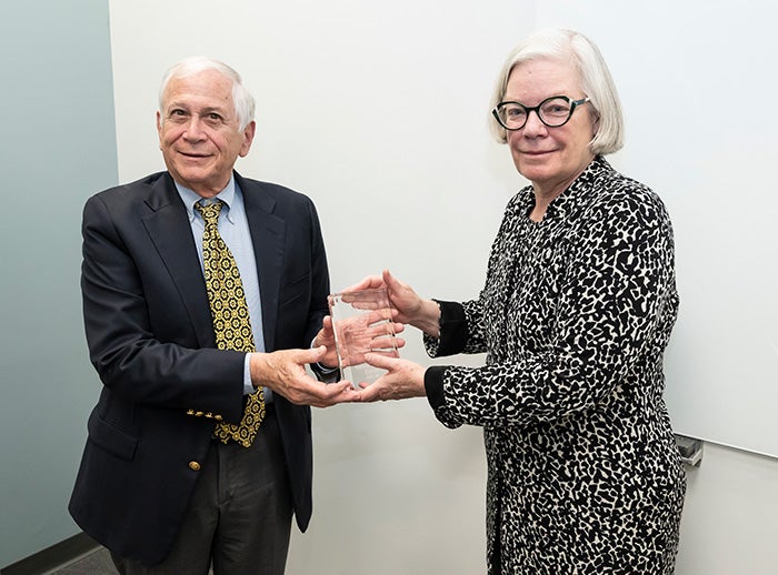 A person in a black and white jacket hands a glass award to a person in a suit and yellow tie