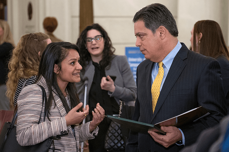 woman of Indian decent in striped blazer talks with man wearing dark suit, blue shirt and yellow tie and holding binder