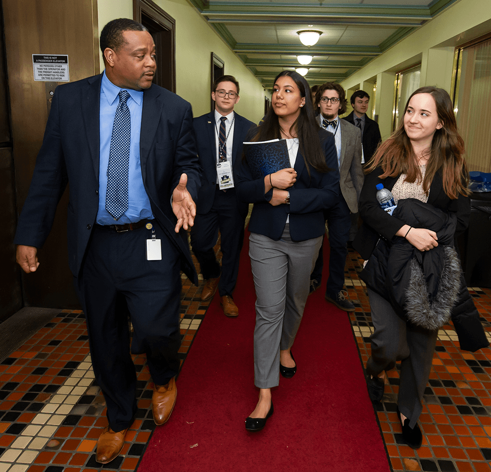 Man in dark blue suit, blue shirt, and patterned tie walks and talks up a hallway with students in business wear. 