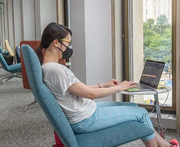 masked student sits in turquoise chair working on laptop and looking out onto Schenley Plaza