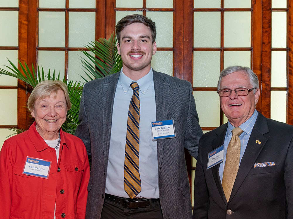 Older white woman with short blond hair, tall young white man with brown hair and mustache and blue/gold suit/tie, and older white man with short gray hair, glasses, navy blue suit, gold tie and script Pitt pin on lapel. Stand in front of windowpane backdrop.