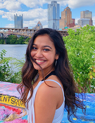 photo of young woman of south Asian descent smiling for camera, standing in front of river, bridge and Pittsburgh cityscape