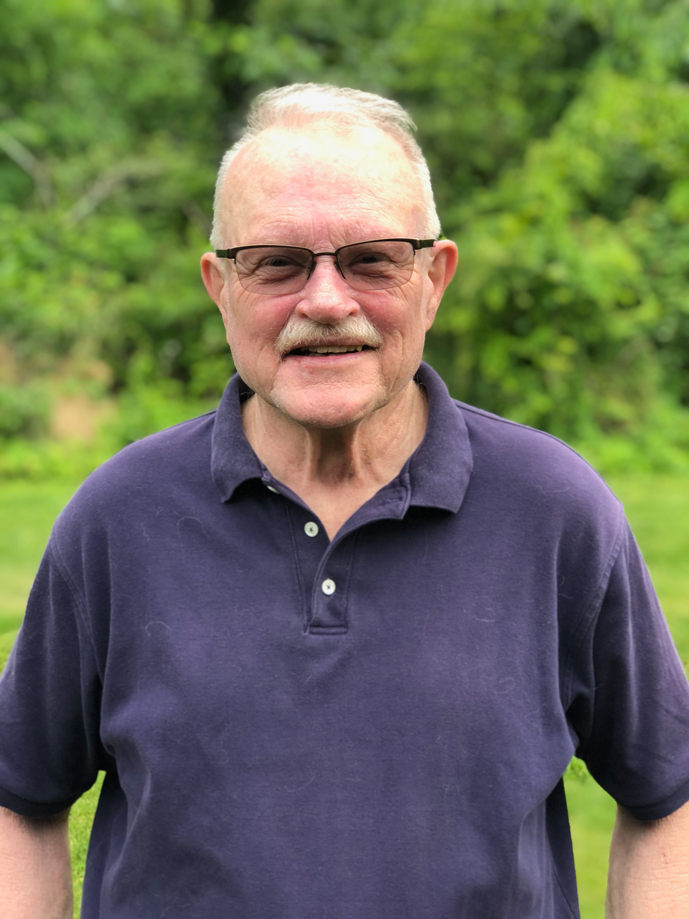 man with light gray hair and mustache wears glasses and purple polo shirt