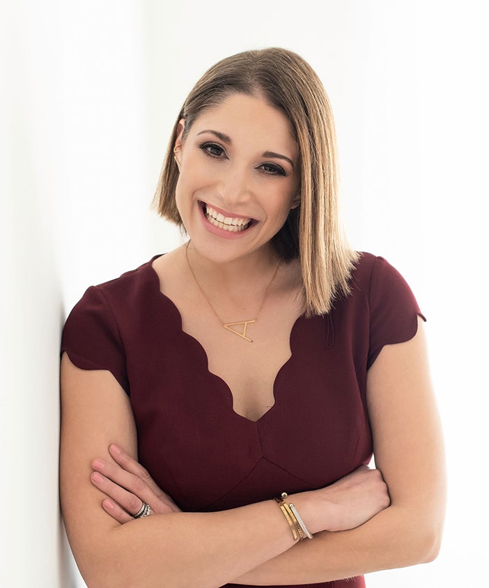 woman wearing plum, scalloped shirt leaning against white wall