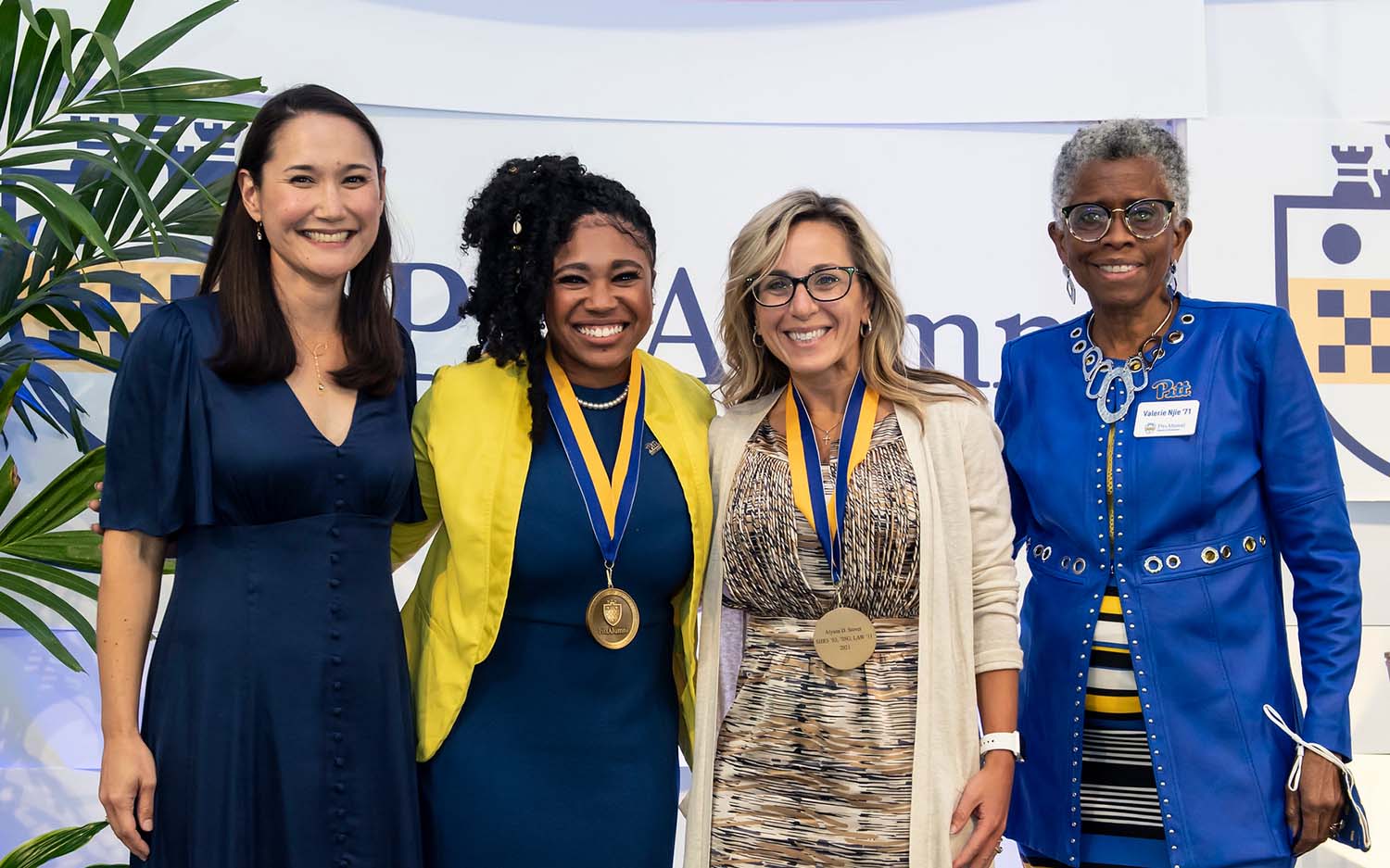 Four women in dark blue and gold clothing pose for photo in front of Pitt seal