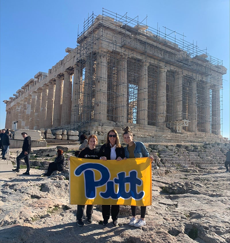Three people hold a yellow Pitt flag in front of ruins