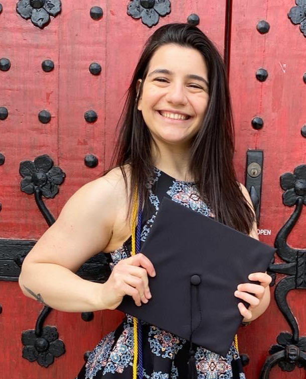 young woman wearing graduation cords holds mortarboard in front of red doors with black hardware