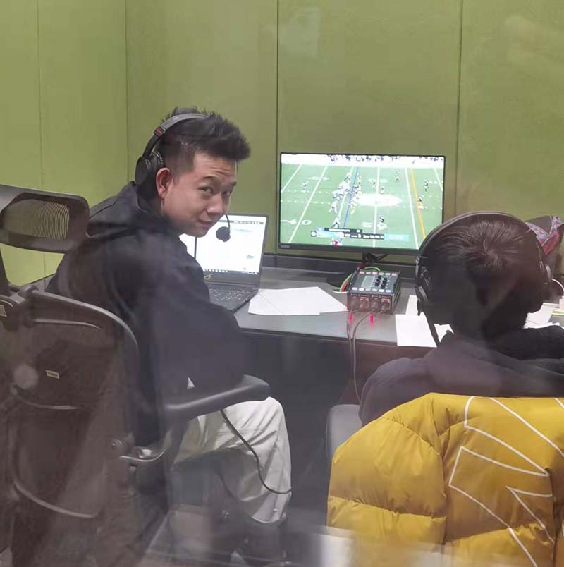 Two men wearing headsets and in rolling chairs sit in front of computer monitor with american football on screen