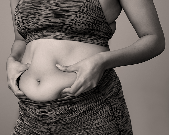 Grayscale photo of woman's torso in sports bra and matching sports pants holds tummy flab with her two hands