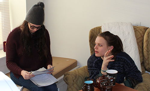 Two young women sit by window, one on window seat and another in wingback chair, looking over papers