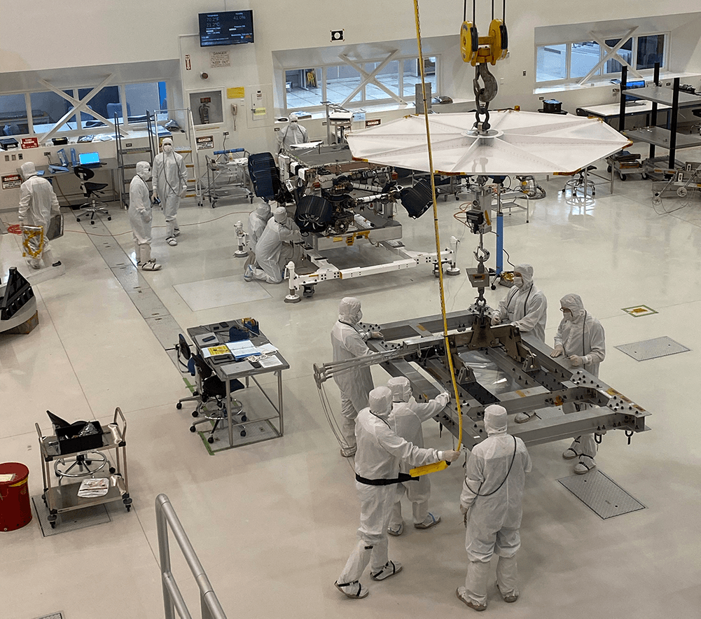 People in white coverall suits, shoe covers, and face masks work on machinery in a large open space.