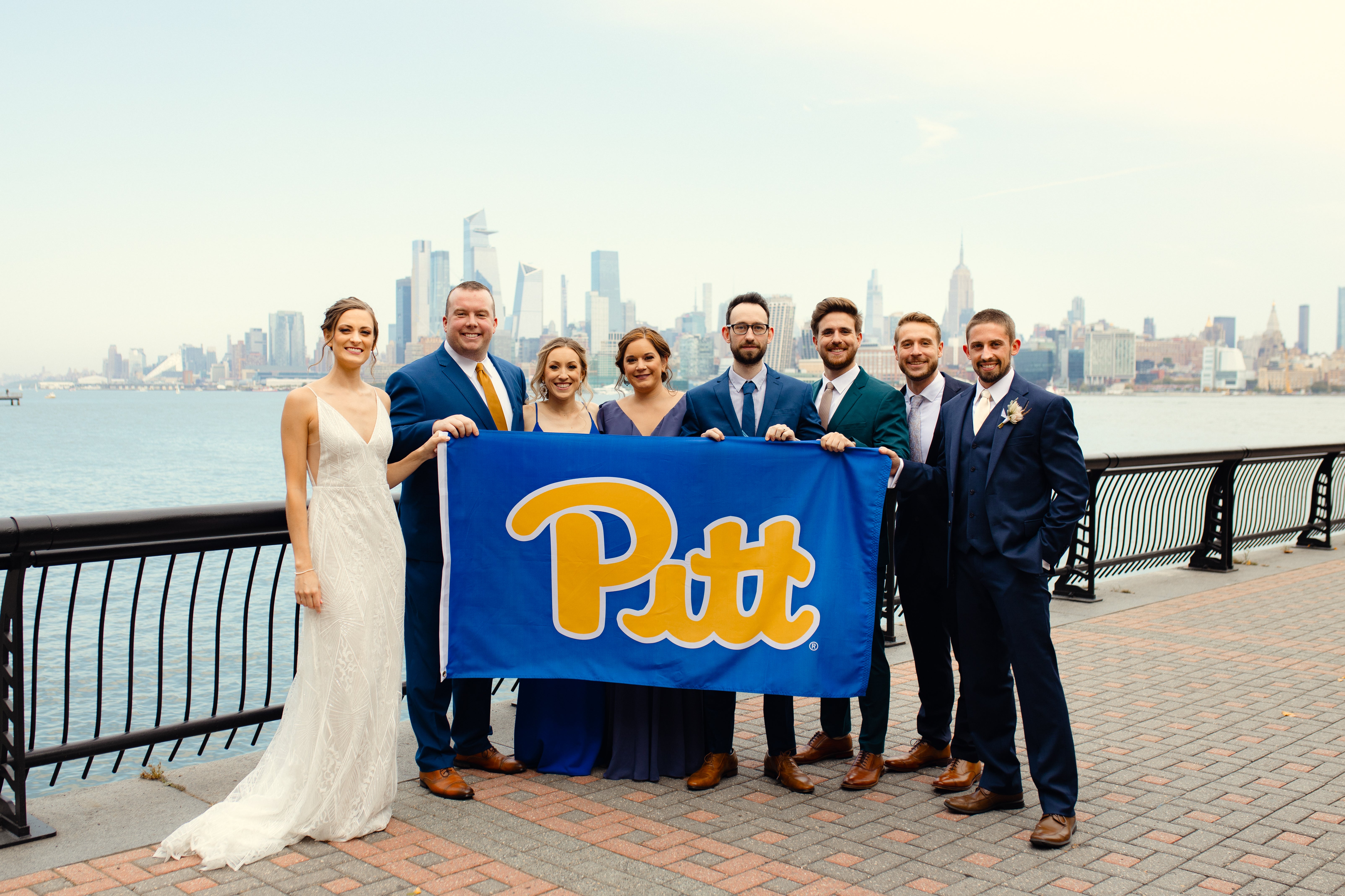 bride in white dress, groom and groomsmen in blue suits, and bridesmaids in blue dresses on Pitt script flag in front of New York City skyline across the Hudson