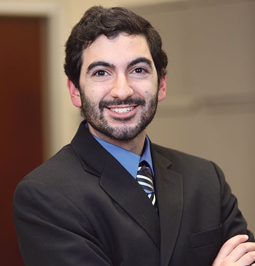 man with dark hair and cropped beard in suit with arms crossed