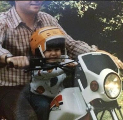 A toddler wears a helmet while riding on her father's lap on a motorcycle