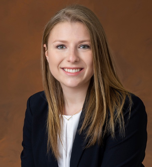 Young woman with long, straight light-brown hair in black blazer and white blouse, smiles at camera while sitting in front of chocolate brown backdrop.