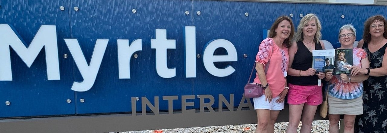 Four women pose with a Pitt Magazine in front of a Myrtle sign