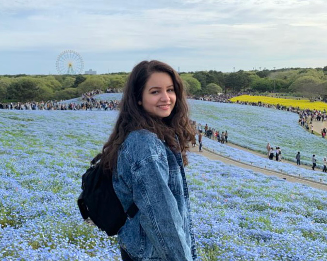 Young woman in denim jacket stands in field of small flowering blue flowers; lines of people, trees and a ferris wheel are visible in the background