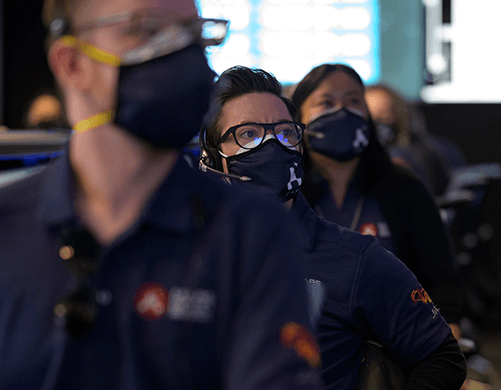 A row of people, in navy blue masks and navy blue polo shirts with the red NASA logo, look up in the same direction, with screens behind them.