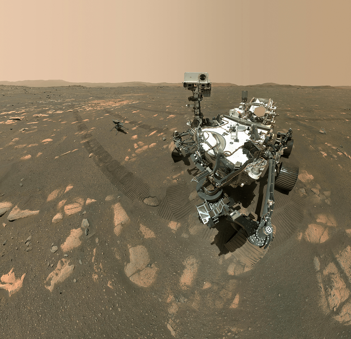 The Perseverance rover and its helicoper appear on the gray-brown floor of Mars, interspersed with orangish rocks. A ridge is visible on the horizon, and the sky is a hazy orange.