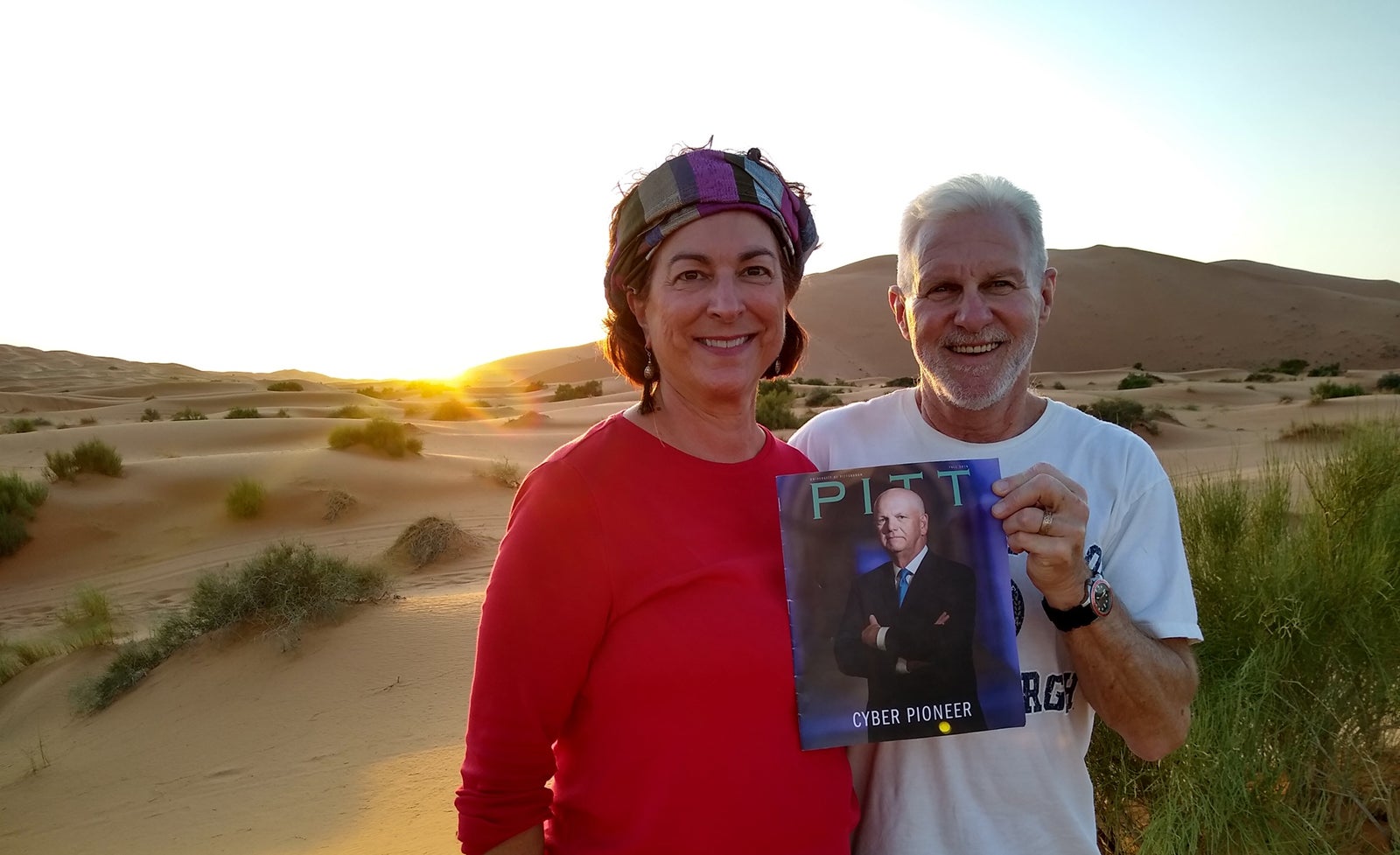 woman in red shirt and headband and man in white University of Pittsburgh T-shirt hold Pitt Magazine Fall 2019 issue as the sun goes down on sandy desert landscape