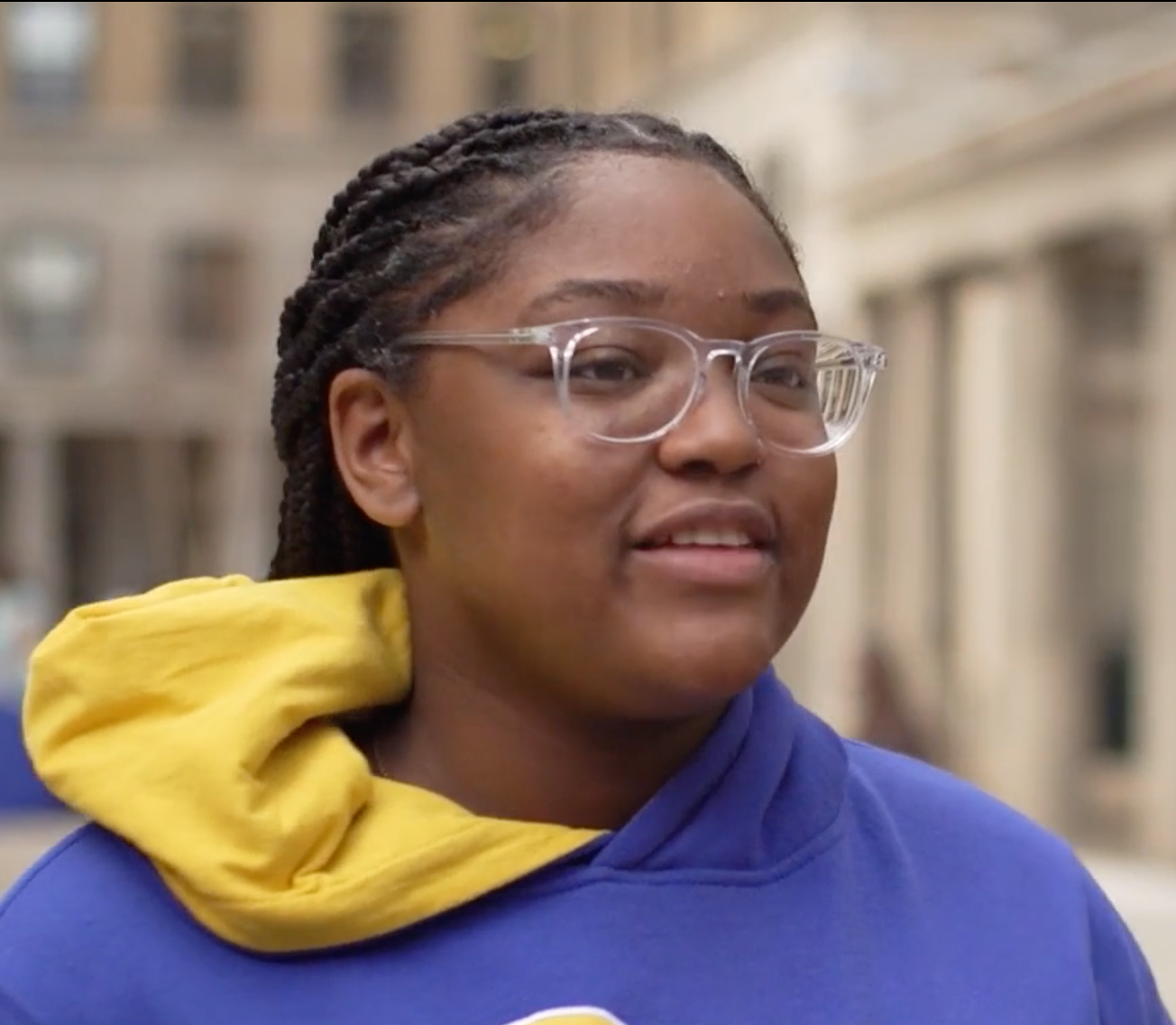 Young Black woman with clear-framed glasses, braids and blue and gold hoodie stands in front of buildings