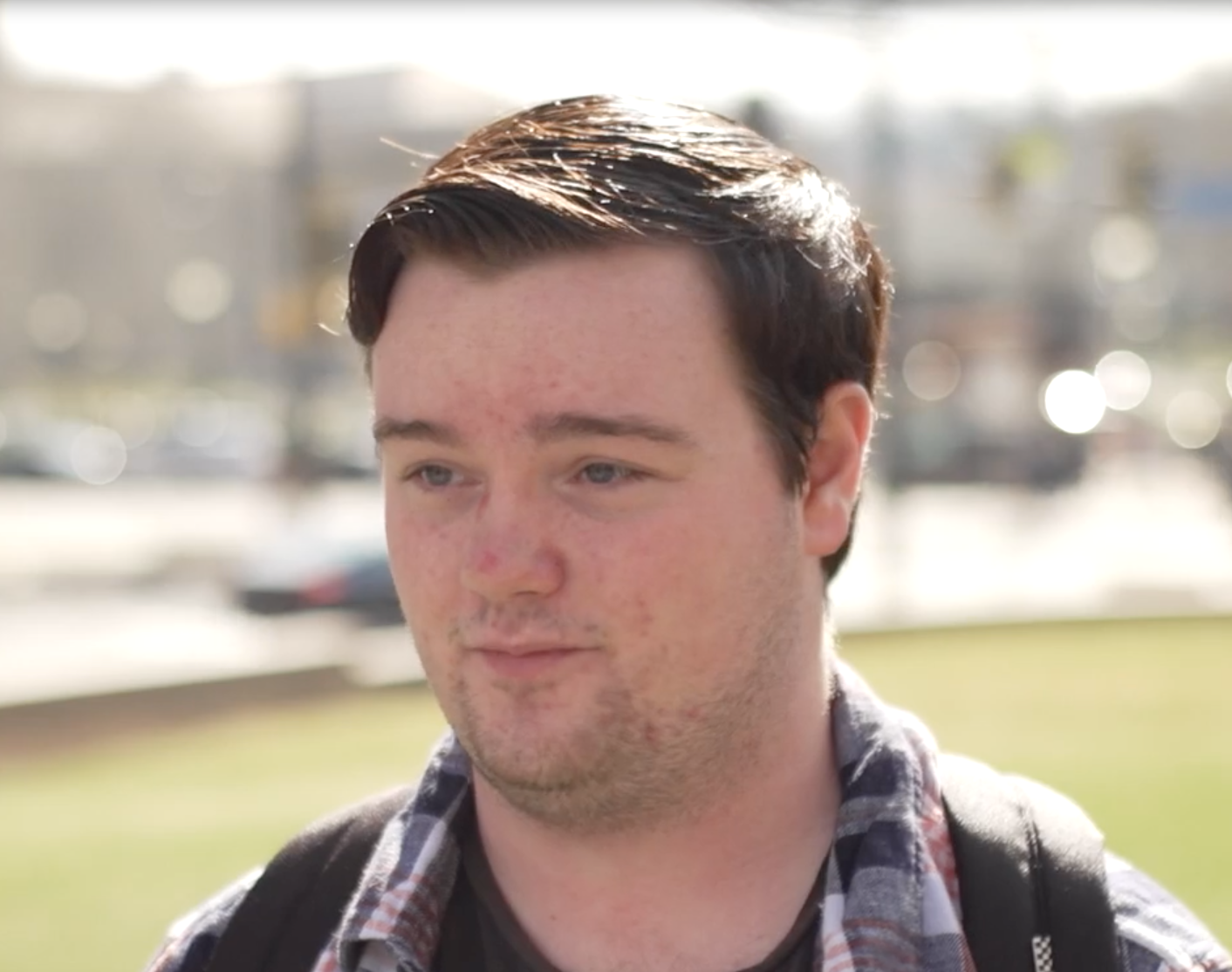 headshot of young white man standing in grass in sunshine, wearing plaid shirt and backpack