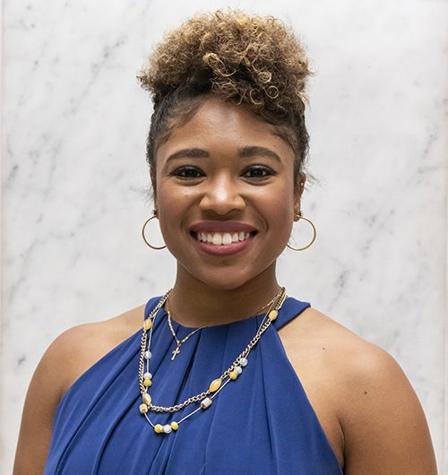 Black woman in blue blouse with gold jewelry standing in front of white marble background