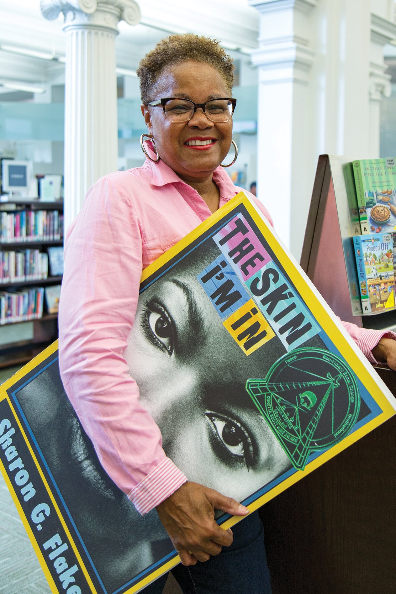 Sharon G. Flake on the first floor of Carnegie Library of Pittsburgh Main's building, holding a blow-up of the cover of The Skin I'm In