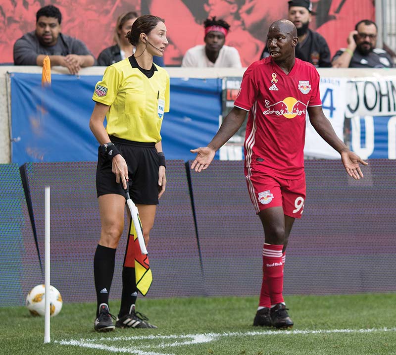 woman in yellow shirt and black shorts holding checkered flag talks to male soccer player in red shirt and shorts at the corner of the pitch