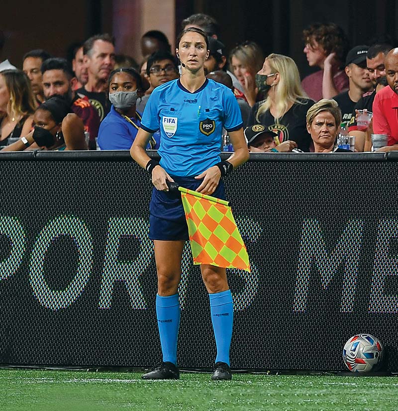 Woman wear "pro" teal ref shirt and black shorts, holds yellow and orange checkered flag on sideline of pitch