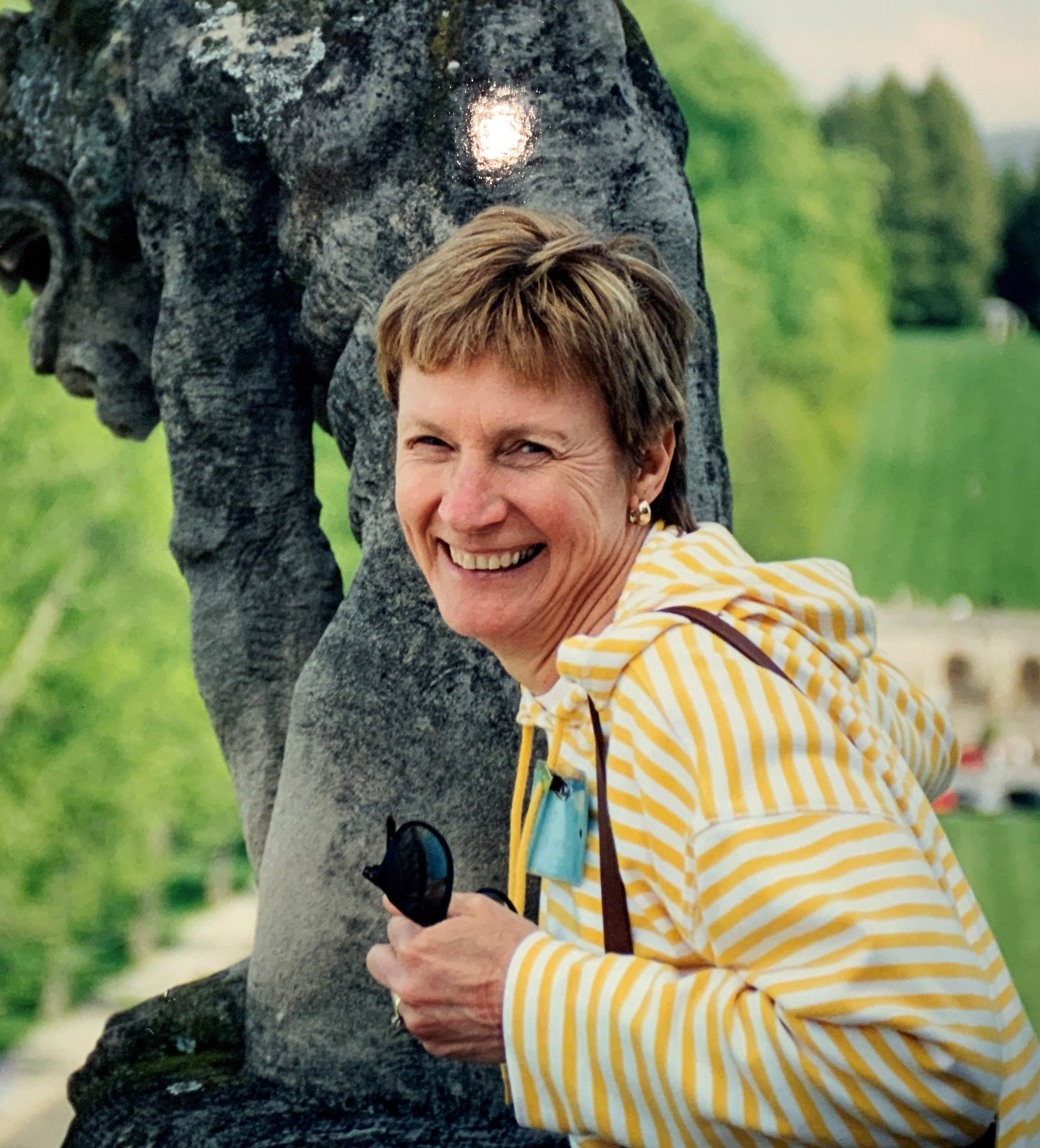 woman with short light brown hair smiles at camera, wearing white and yellow striped hoodie and standing in front of dark-stone sculpture