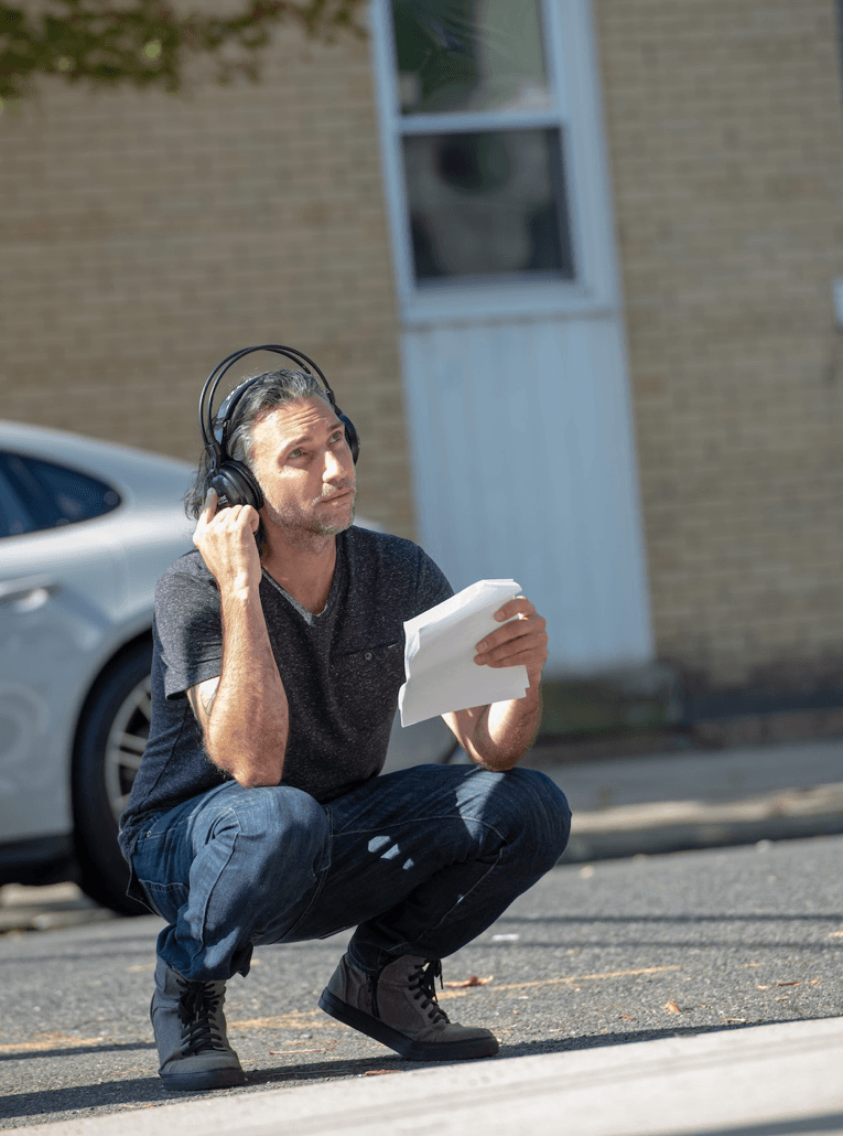 Man with shoulder-length silver hair, wearing jeans and dark t-shirt, crouches on pavement and holds earphones on head while looking in front of/above him