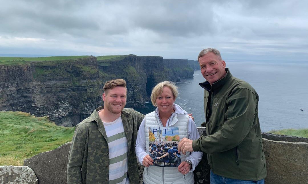 young man with his mother and father, dressed in greens and pale blue-grays, stand with Pitt Magazine Winter 2019 issue in front of the Cliffs of Moher