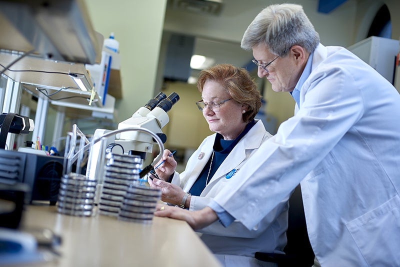 Stout works with tools in a lab while another scientist in a white coat watches over her shoulder