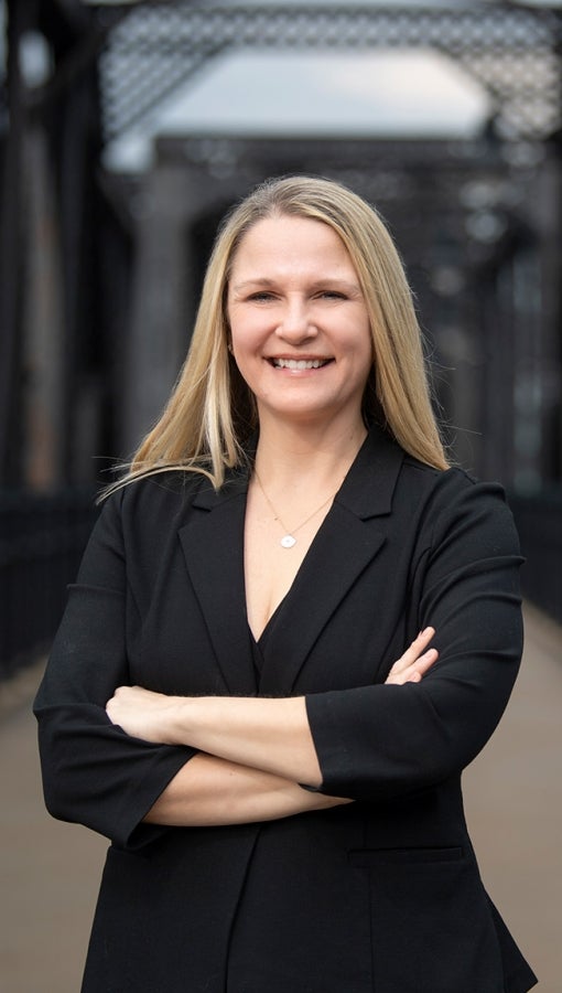 Woman in black blazer and long blond hair smiles at camera and stands on a black, industrial-style bridge