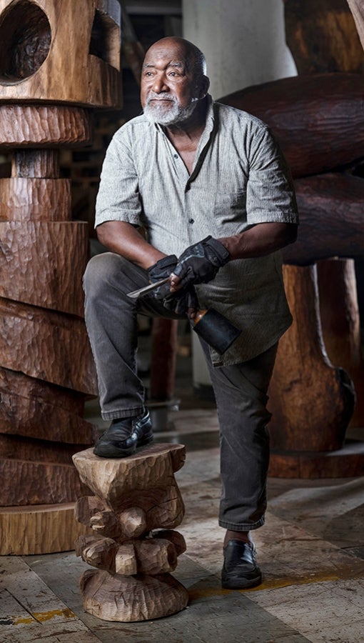 Thaddeus Mosley perches one leg on a wood slab while surrounded by cherry and walnut sculptures