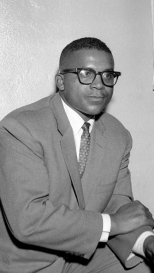 Young black man in suit and tie leans on knee, leg propped up, wears dark rimmed glasses