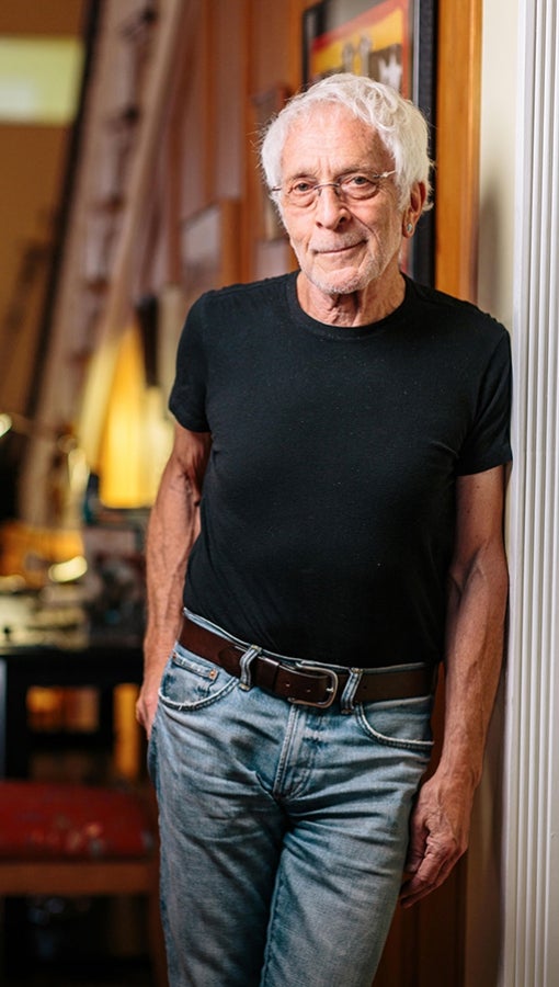 Lee Gutkind, in short-sleeved black t-shirt and jeans, stands in hallway of house, framed images and table lamp visible in background
