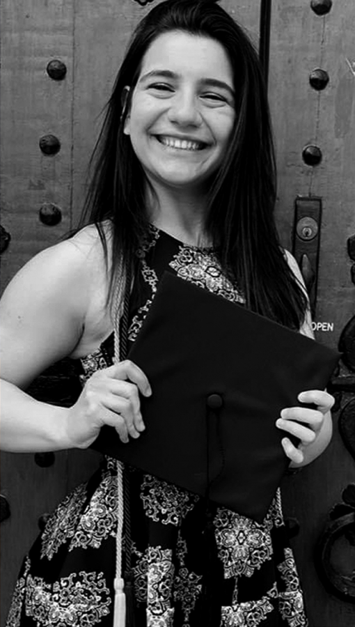 black and white image of young woman wearing graduation cords holds mortarboard in front of doors with ornate hardware