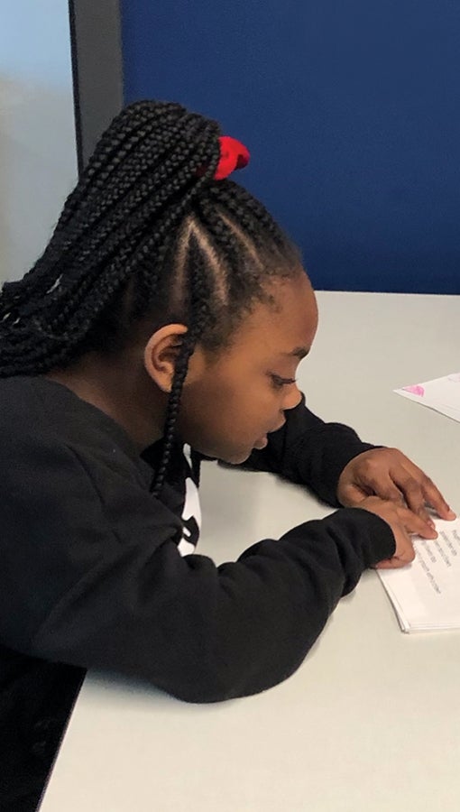 a little girl, her hair braided and pulled up in a scrunchy, practices reading, following the lines with her finger